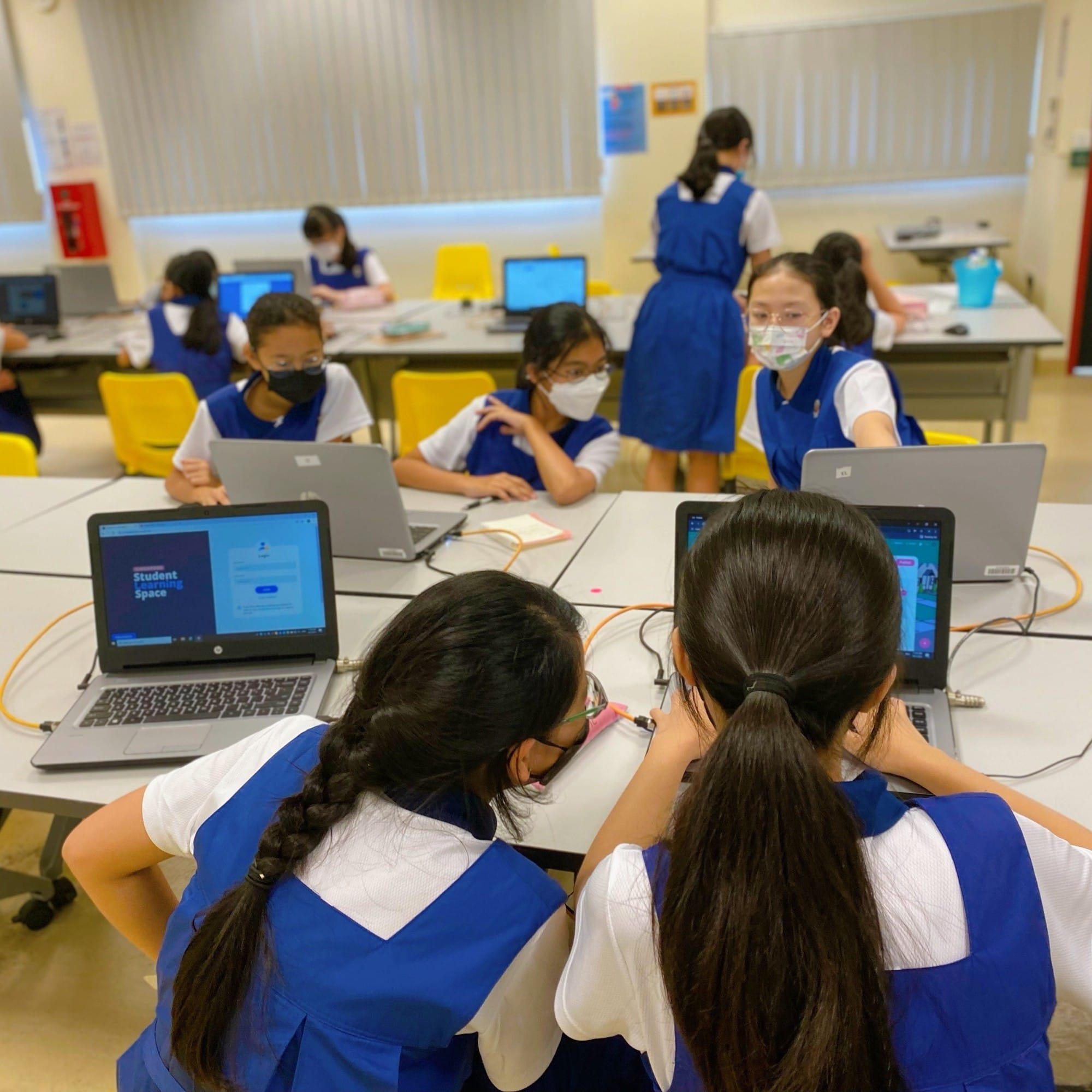 Girls in blue school uniforms and masks work on laptops at tables in a classroom computer lab.